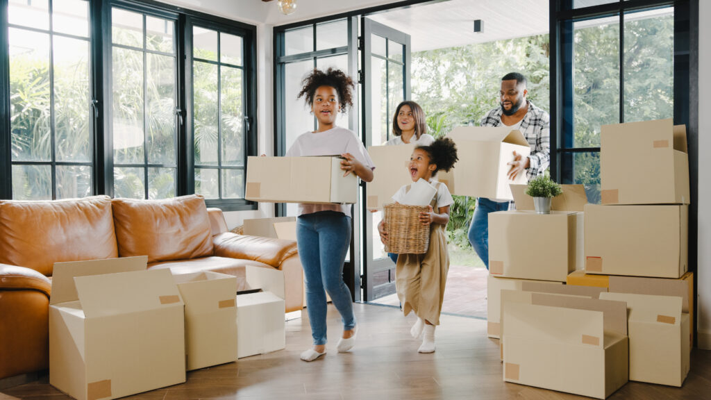 Happy African American young family bought new house. Mom, Dad, and child smiling happy hold cardboard boxes for move object walking into big modern home. New real estate dwelling, loan and mortgage.